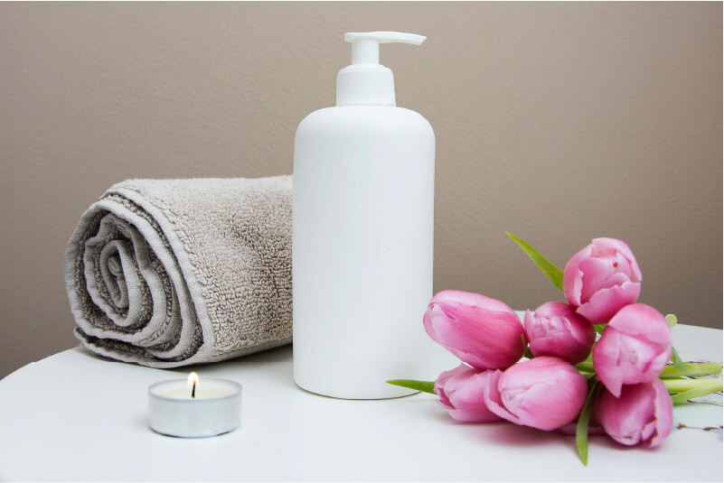 Image of lotion with a candle and tan towel to the left, and pink tulips to the right