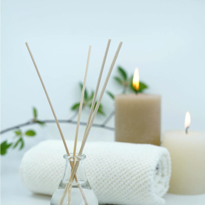 Image composition of small sticks in an essential oil diffuser in front of a white towel, a lit tall and short tan candle, and a fresh branch with green leaves